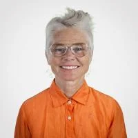 Zoe Welch facing the camera head-on, smiling, with upswept silver hair, wearing an orange shirt.