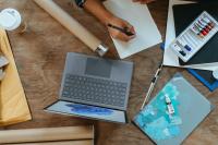 Image from above of a work surface with a laptop, box of paints, and someone's hand writing ona a sheet of paper